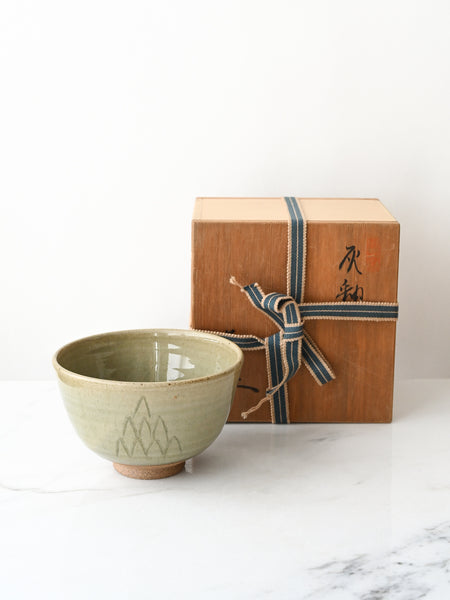 Green ceramic bowl in front of a wooden gift box with a ribbon on a white marble surface.